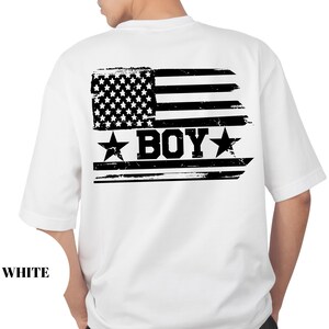 Puede incluir: Camiseta blanca con un dise&ntilde;o de bandera americana desgastada en la espalda. El gr&aacute;fico de la bandera incluye estrellas, rayas y la palabra "BOY" en negrita, con estrellas a cada lado. La palabra "WHITE" est&aacute; impresa en la parte inferior.