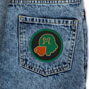 Puede incluir: Vaqueros azul claro con un parche circular en el bolsillo trasero. El parche presenta una figura de dibujos animados verde con gafas sosteniendo una taza de café marrón, sobre un fondo negro y verde.