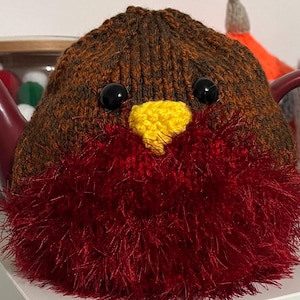 May include: A ceramic teapot with a knitted robin tea cosy. The cosy is brown and red with black eyes and a yellow beak. The teapot has a red handle and spout. The cosy is designed to look like a robin bird.