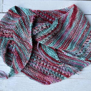 May include: A triangular knit shawl featuring a variegated color pattern of teal, magenta, and soft pink. The garment is constructed with a combination of garter stitch and openwork sections, finished with small tassels at the pointed ends.