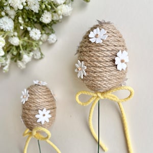 May include: Two brown twine Easter eggs decorated with white paper flowers and yellow ribbon bows on green stems.
