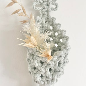 May include: A pale grey macrame plant holder with a woven basket design, featuring a decorative tassel at the bottom and a floral arrangement of dried beige and white flowers. The holder is suspended against a white background.
