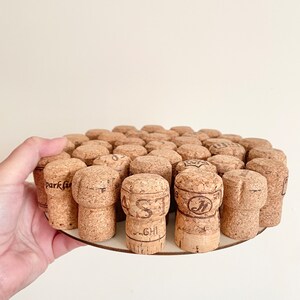 May include: A round wooden coaster with a circle of cork bottle stoppers. The stoppers are arranged in a circular pattern and are all different sizes and shapes. The coaster is a light brown colour and has a smooth surface.