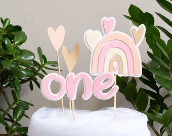 May include: Pink and gold cake topper with the word "one" and a rainbow design. The topper is on a white cake.