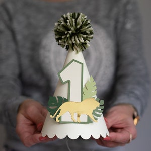 May include: A white paper party hat with a green and white pom-pom on top. The hat has a number "1" on the front, a gold lion, and green leaves.