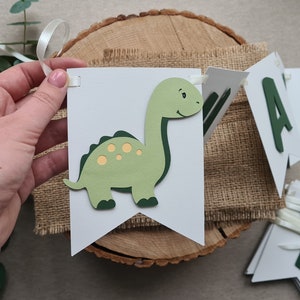 May include: A green dinosaur banner with the word "Happy" in green letters. The banner is made of white cardstock and has a burlap background.
