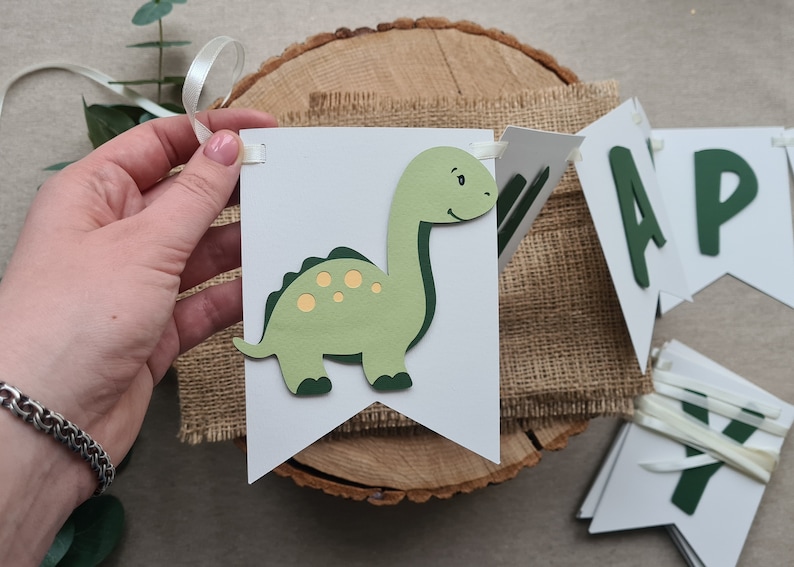 May include: A green dinosaur banner with the word "Happy" in green letters. The banner is made of white cardstock and has a burlap background.