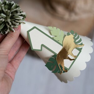 May include: A white paper party hat with a green pom-pom on top. The hat has a green number one and a gold lion cutout on the front.