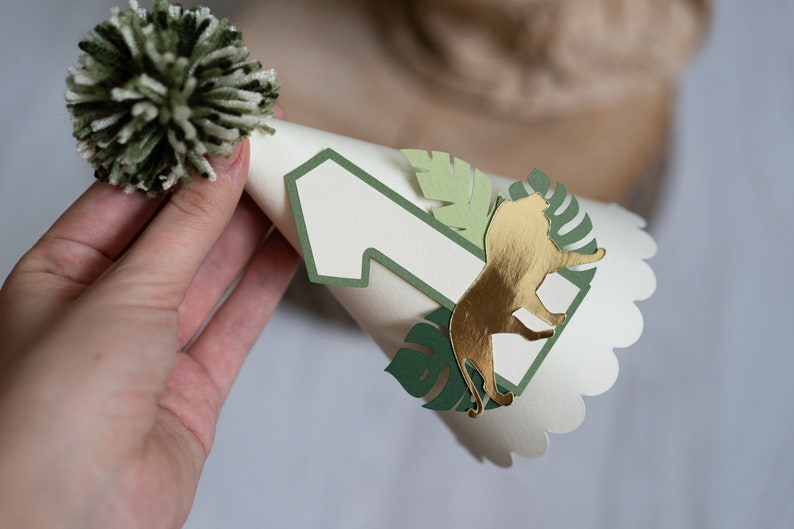May include: A white paper party hat with a green pom-pom on top. The hat has a green number one and a gold lion cutout on the front.