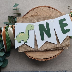 May include: A green dinosaur and the word "ONE" in green letters on a white banner with burlap fabric behind it. The banner is on a wooden slice with green and brown ribbons.