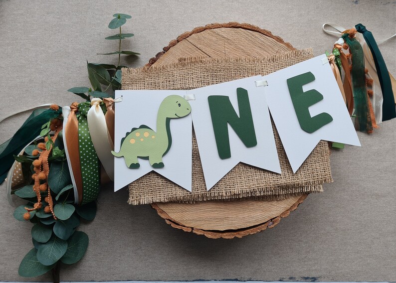 May include: A green dinosaur and the word "ONE" in green letters on a white banner with burlap fabric behind it. The banner is on a wooden slice with green and brown ribbons.
