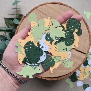 May include: A collection of green, yellow, and white paper cutouts of dinosaurs and monstera leaves. The cutouts are scattered on a wooden surface.