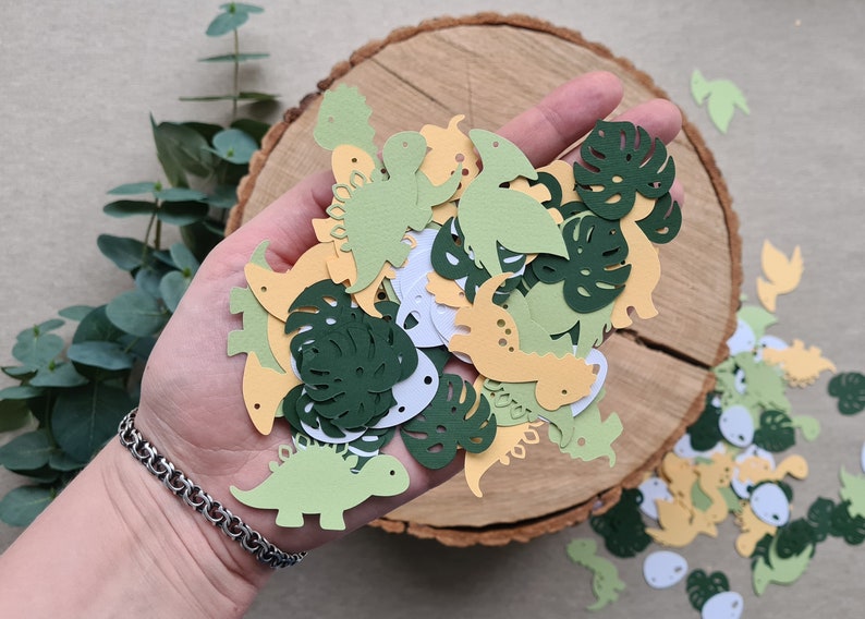 May include: A collection of green, yellow, and white paper cutouts of dinosaurs and monstera leaves. The cutouts are scattered on a wooden surface.