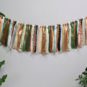 May include: A green, white, and gold fabric banner with a variety of textures and patterns. The banner is strung with a white cord and has small pom-poms throughout.