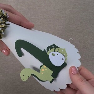 May include: A white paper party hat with a green dinosaur design. The dinosaur has a green body with yellow spots, a green head with a white eye, and a green leaf on its back. The hat has a white pom-pom on top.