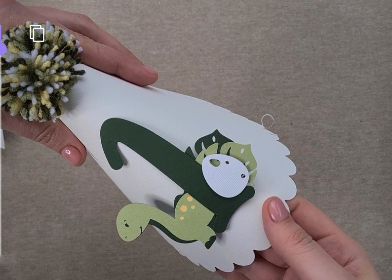 May include: A white paper party hat with a green dinosaur design. The dinosaur has a green body with yellow spots, a green head with a white eye, and a green leaf on its back. The hat has a white pom-pom on top.