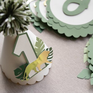 May include: A white paper party hat with a green pom-pom on top. The hat has a green number one, a gold lion, and green leaves on it.
