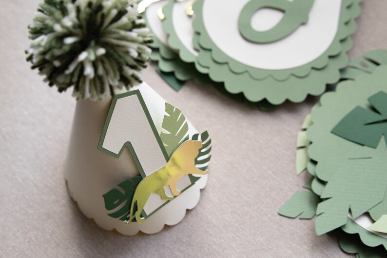 May include: A white paper party hat with a green pom-pom on top. The hat has a green number one, a gold lion, and green leaves on it.