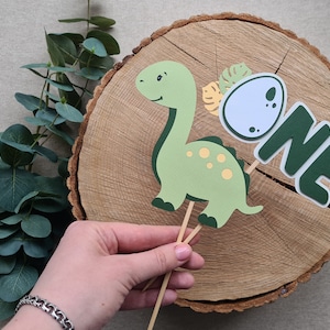 May include: Green dinosaur cake topper with a yellow egg and the word "ONE" in green letters. The topper is on a wooden slice.