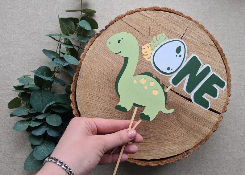 May include: Green dinosaur cake topper with a yellow egg and the word "ONE" in green letters. The topper is on a wooden slice.