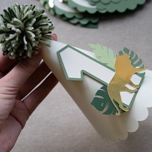 May include: A white party hat with a green and gold jungle theme. The hat has a number one, a lion, and leaves cut out of paper. A white pom-pom sits on top of the hat.