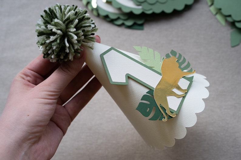 May include: A white party hat with a green and gold jungle theme. The hat has a number one, a lion, and leaves cut out of paper. A white pom-pom sits on top of the hat.