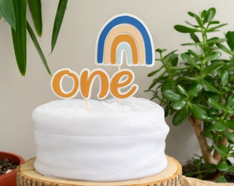 May include: A white cake with a blue, orange, and white rainbow cake topper and a brown "one" cake topper.