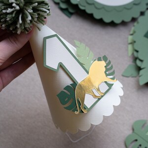 May include: A white paper party hat with a green pom-pom on top. The hat has a green and gold lion cutout, a green leaf cutout, and a white number one.