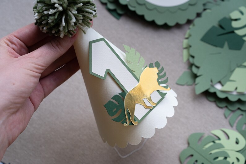 May include: A white paper party hat with a green pom-pom on top. The hat has a green and gold lion cutout, a green leaf cutout, and a white number one.