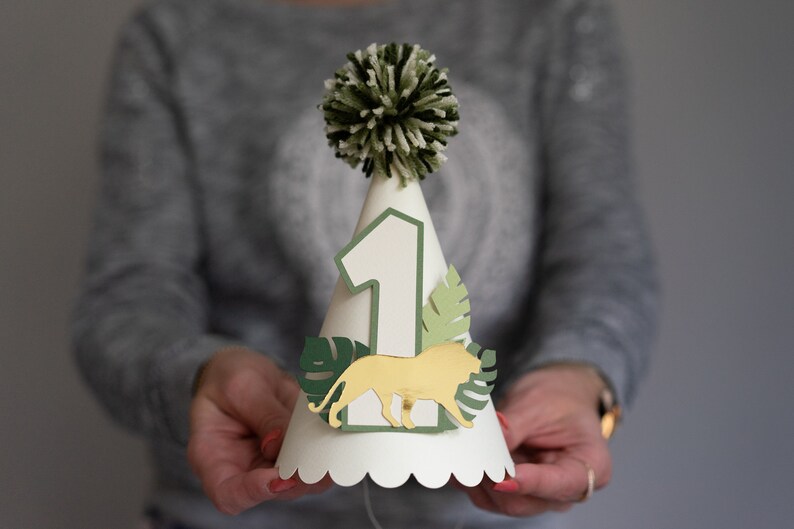 May include: A white paper party hat with a green and white pom-pom on top. The hat has a number "1" on the front, a gold lion, and green leaves.
