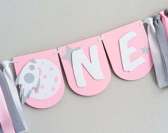 May include: Pink and gray banner with the word "ONE" in white letters, a rocket and moon, and stars. The banner has pink and gray ribbon streamers.
