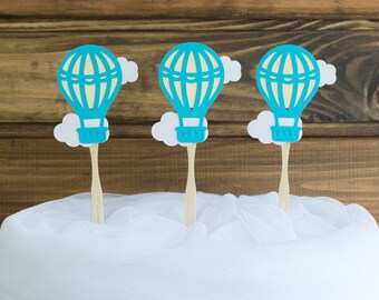 May include: Three blue hot air balloon cake toppers with white clouds, each on a wooden stick. The toppers are on a white cake covered in white tulle.