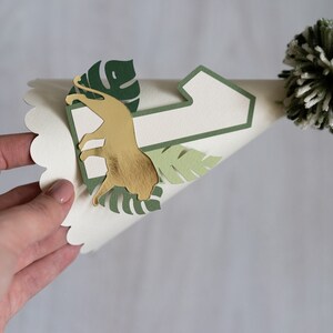 May include: A white paper party hat with a green number one, a gold lion cutout, and green leaves. The hat has a white and green pom-pom on top.