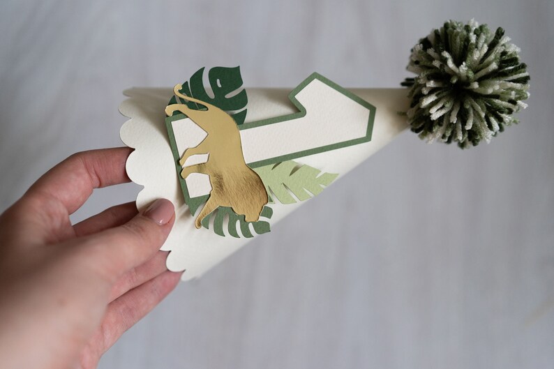 May include: A white paper party hat with a green number one, a gold lion cutout, and green leaves. The hat has a white and green pom-pom on top.