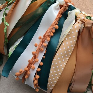 May include: A collection of ribbons in various colors and patterns, including white, cream, brown, green, and a ribbon with white polka dots on a brown background. Some ribbons have a fringe of orange pom-poms.