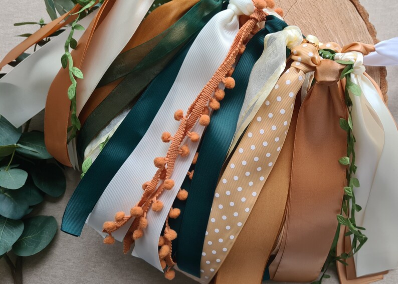 May include: A collection of ribbons in various colors and patterns, including white, cream, brown, green, and a ribbon with white polka dots on a brown background. Some ribbons have a fringe of orange pom-poms.