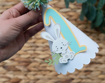 May include: A white paper party hat with a scalloped edge. The hat has a blue and yellow number one on it and a white bunny with green leaves.