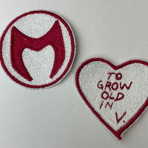 Puede incluir: Dos parches bordados, uno circular con un símbolo "M" rojo y el otro en forma de corazón con el texto "To grow old in V."