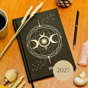 May include: A black hardback planner featuring a gold foil celestial design with a triple moon symbol and the year 2027 printed on the cover. The planner is accompanied by a black pen, a light pink crystal, a seashell, and dried wheat stalks.