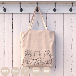 May include: A beige tote bag with three cats wearing Christmas hats and lights. The cats are outlined in black and are sitting in a row. The bag is hanging on a white wooden wall with hooks.