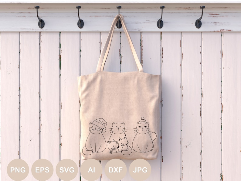 May include: A beige tote bag with three cats wearing Christmas hats and lights. The cats are outlined in black and are sitting in a row. The bag is hanging on a white wooden wall with hooks.