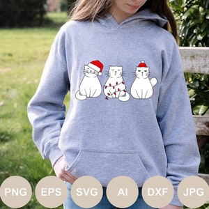 May include: A gray hoodie with a graphic of three white cats wearing Santa hats. The cats are in a row, with the middle cat wrapped in Christmas lights.