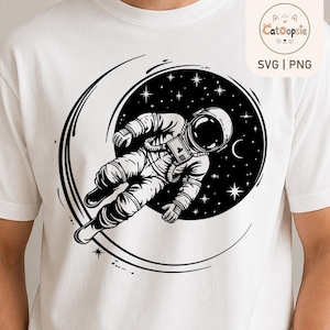 May include: A white short-sleeved t-shirt featuring a black graphic print of an astronaut floating in space within a circular frame. The design includes stars and a crescent moon set against a solid black background, creating a high-contrast look.