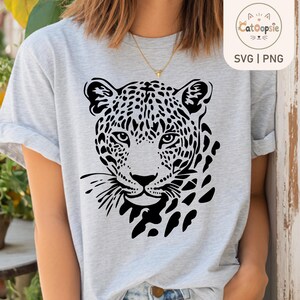 May include: A light gray heathered t-shirt featuring a bold black graphic print of a leopard face on the front. The leopard design displays detailed spots, whiskers, and piercing eyes. The shirt has short sleeves and a classic crew neckline.