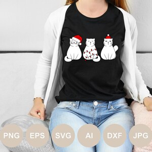 May include: A black t-shirt with a white design of three cats wearing Santa hats. The cat in the middle is tangled in Christmas lights.