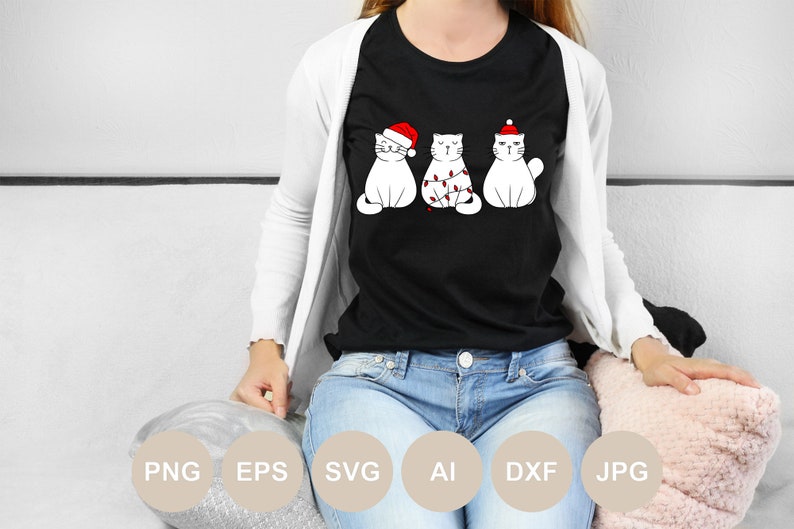 May include: A black t-shirt with a white design of three cats wearing Santa hats. The cat in the middle is tangled in Christmas lights.
