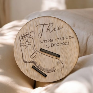May include: A circular wooden plaque engraved with the name "Theo" in script. It displays a baby footprint and birth details: 6:32 PM, 7 lbs 2 oz, and 15 December 2023. Two black bars are positioned below the footprint.