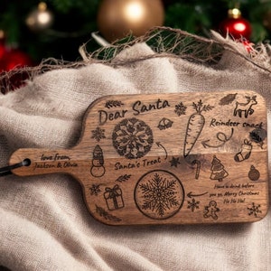 May include: A wooden cutting board with a handle, engraved with Christmas-themed illustrations and text. The text includes "Dear Santa," "Reindeer snack," and "Santa's Treat." The board also features a snowflake, snowman, and gift box designs.