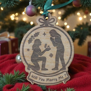 Może przedstawiać: Drewniana ozdoba choinkowa z sceną oświadczyn. Ozdoba przedstawia dwie postacie, jedną klęczącą i prezentującą pierścionek, drugą ze splecionymi dłońmi. Tekst "Will You Marry Me?" jest wygrawerowany na dole.