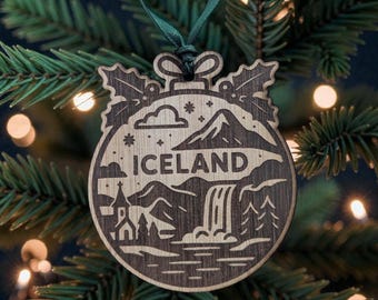 May include: Wooden Christmas bauble with the word "ICELAND" etched into the design. The bauble features a landscape with mountains, a waterfall, and a church. It hangs from a green ribbon on a Christmas tree.